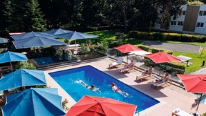 Una piscina con sillas de descanso y sombrillas, rodeada de follaje y un edificio.