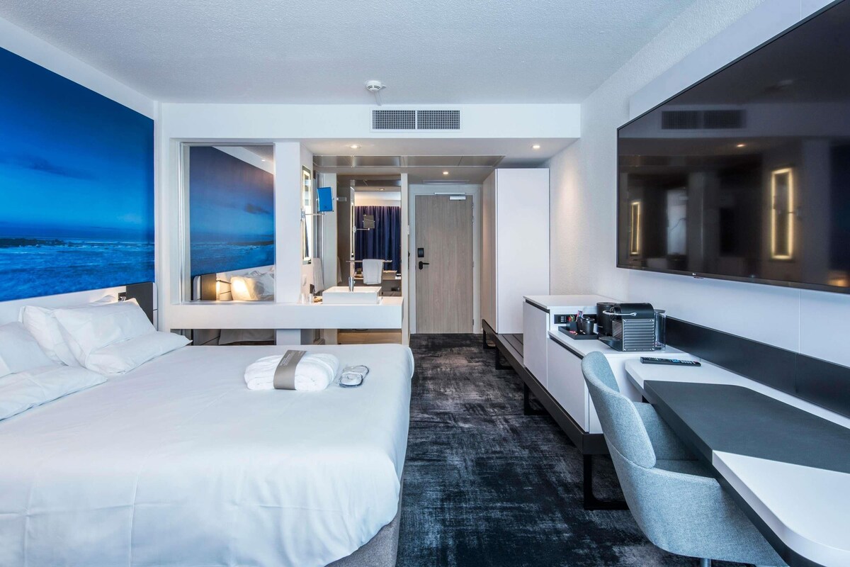 Une chambre d’hôtel moderne équipée d’un lit, d’un bureau et d’un coin salon. La fenêtre offre une vue sur l’océan.