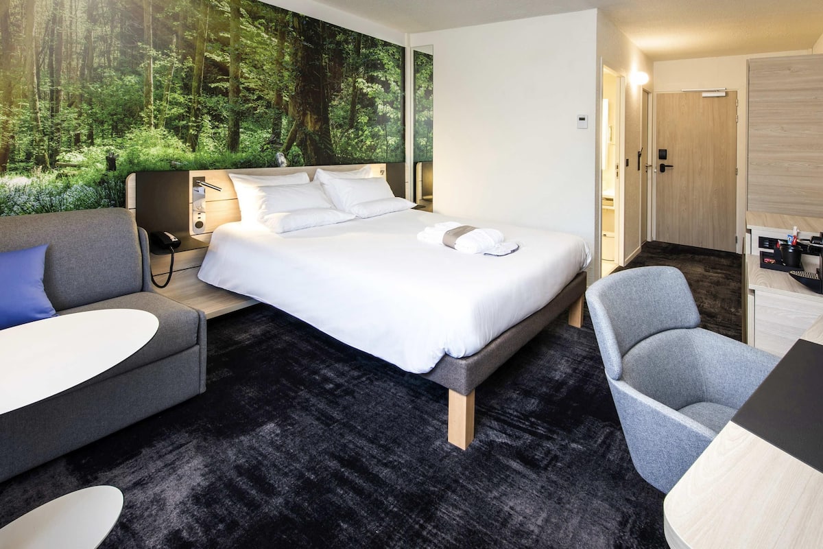 A modern hotel room with a large bed, a sofa, a chair, and a desk.