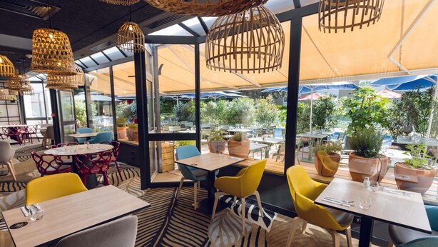 Un comedor interior con mesas y sillas, ventanas amplias y lámparas de mimbre colgantes.
