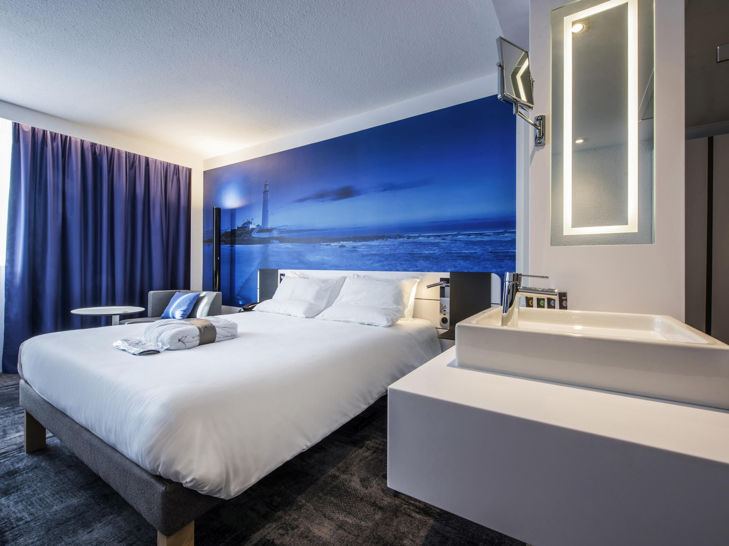 A hotel room with a large bed, a bedside table, a bathroom with a sink, and a wall mural of a lighthouse.