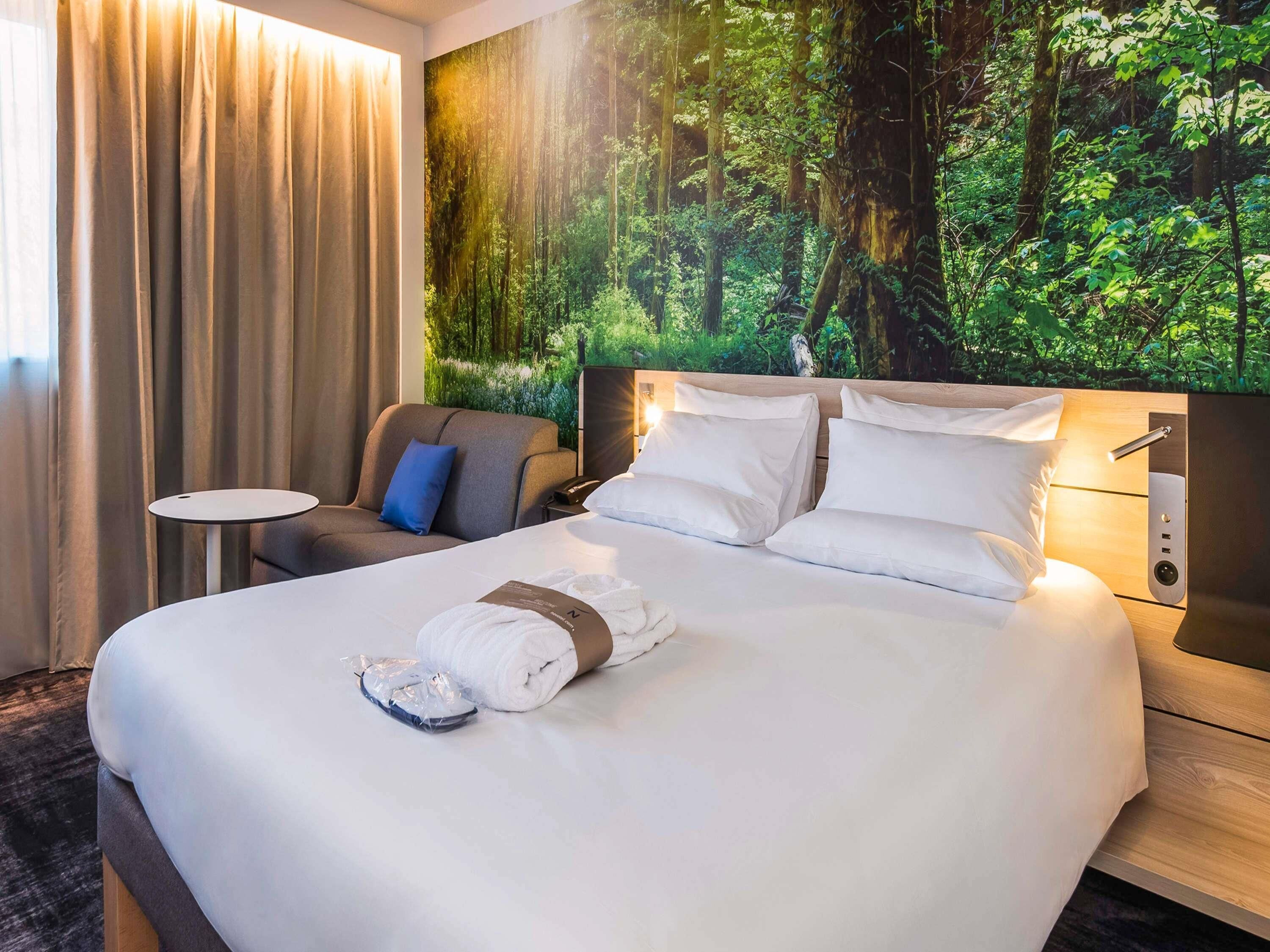 Een hotelkamer met een groot bed, een klein tafeltje, een stoel en een wanddecoratie met een bosmotief.