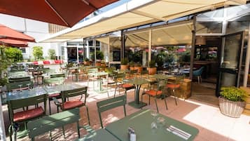 Um café ao ar livre com mesas e cadeiras, protegido por grandes guarda-sóis, e uma área envidraçada com assentos.