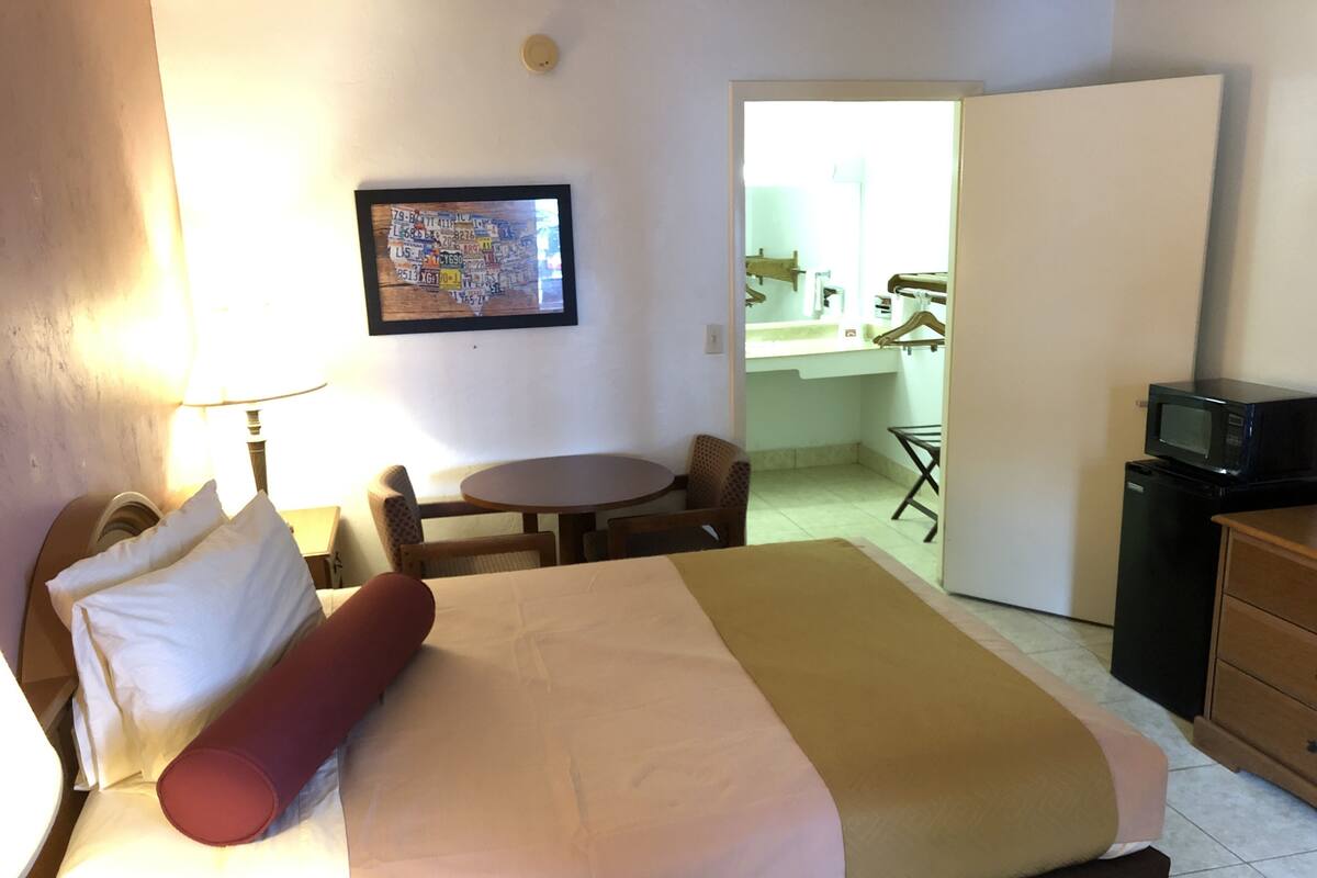 A hotel room with a bed, a desk, a chair, a small table, a microwave, and a bathroom visible through an open door.