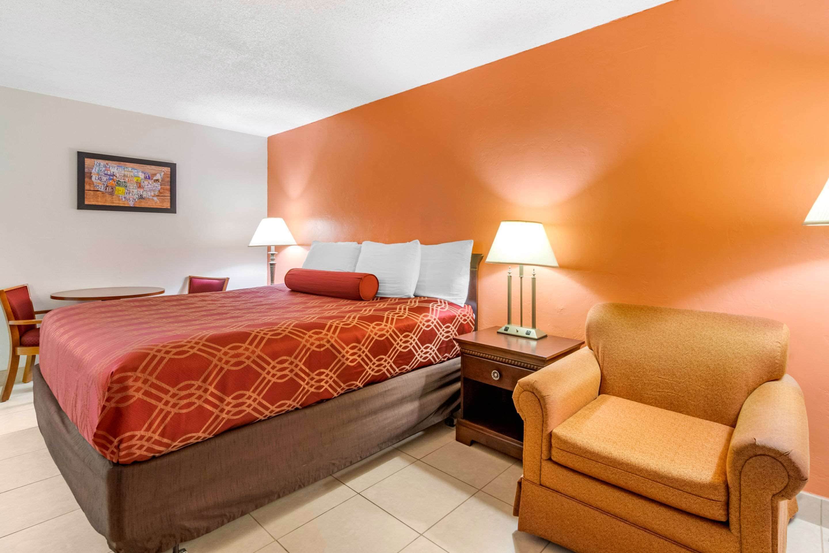 A hotel room with a bed, two bedside tables with lamps, a chair, a small table, and a wall-mounted artwork.