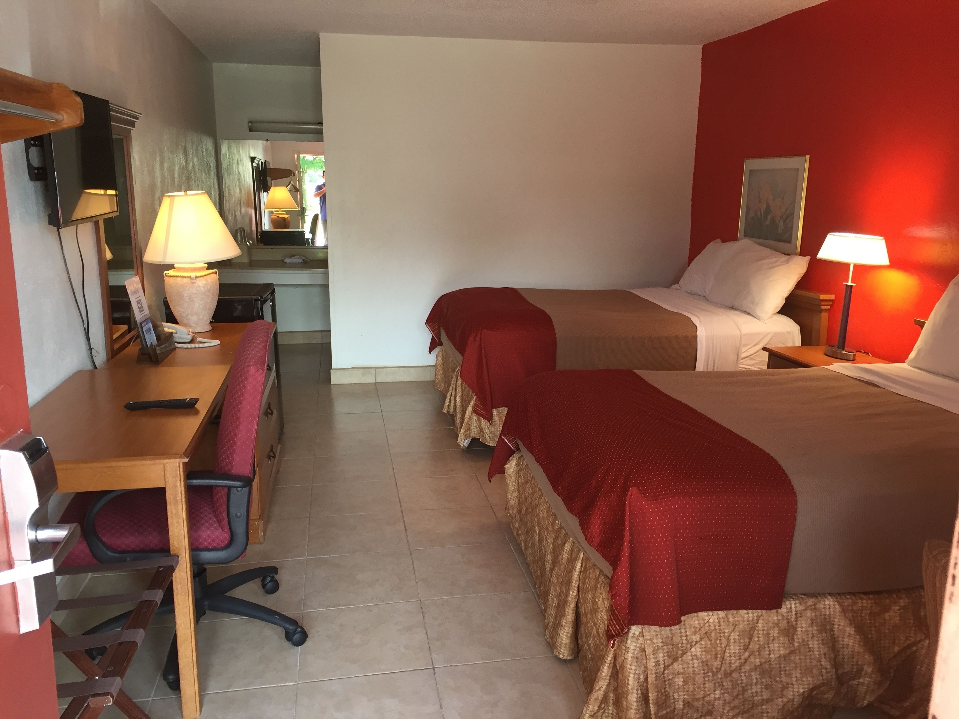 A hotel room with two beds, a desk, a chair, a lamp, and a small kitchenette.