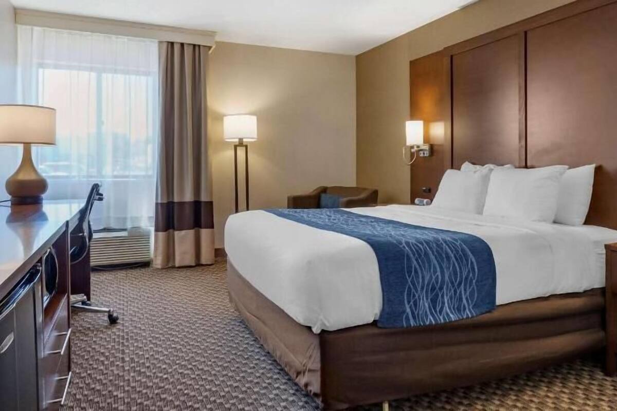 A hotel room with a large bed, bedside lamps, a desk with a telephone, and a view of the city through the window.