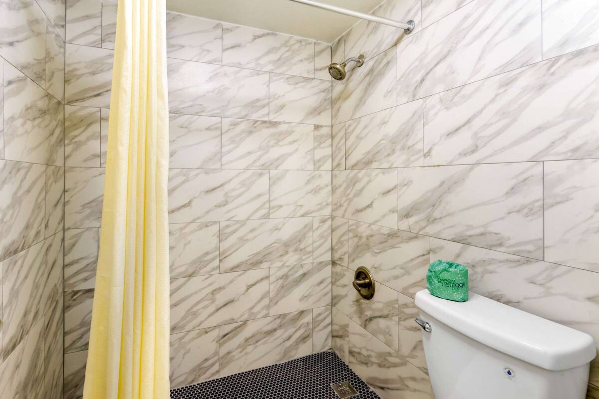 A bathroom with marble walls, a shower curtain, a toilet, and a green bag on the toilet tank.