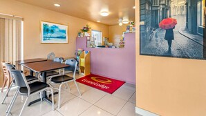 Una hall d'albergo con una reception, una sala da pranzo con tavolo e sedie, un quadro appeso al muro e un tappeto rosso con il logo dell'hotel.