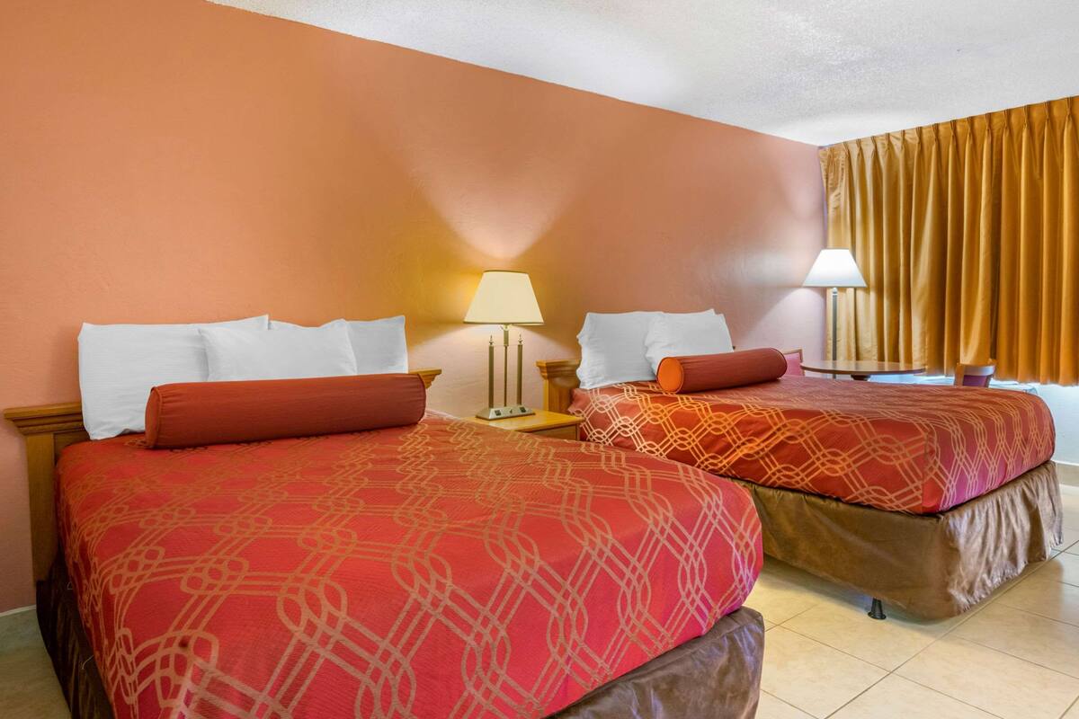 A hotel room with two beds, each with a red patterned bedspread and white pillows. There are two bedside tables with lamps.