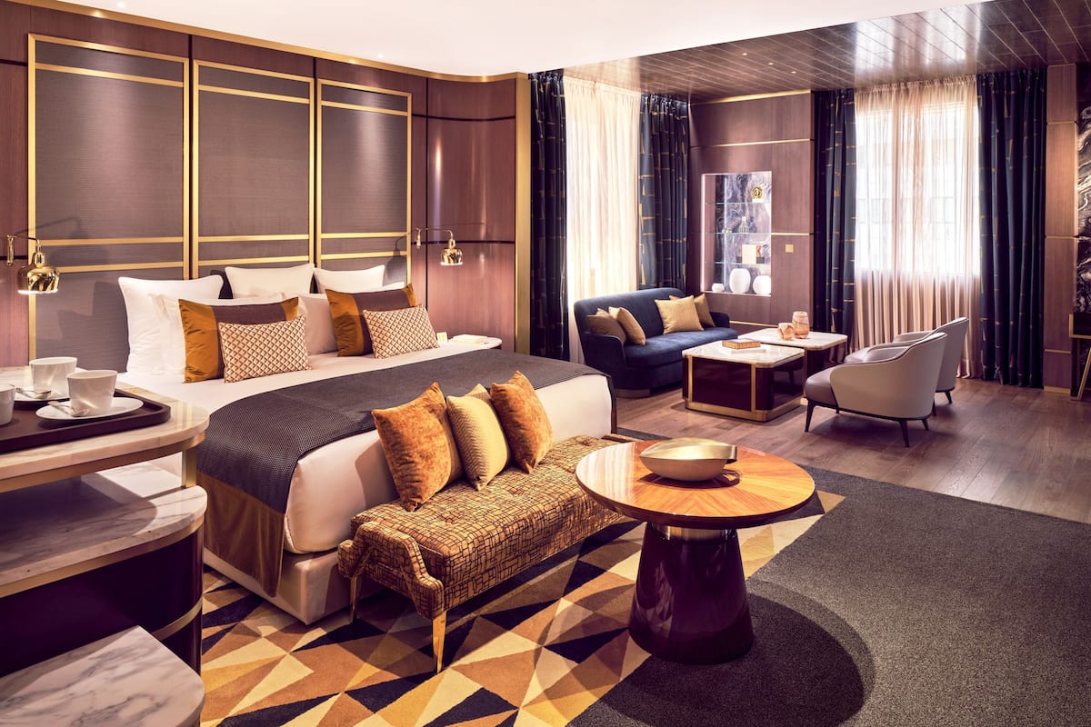 A modern hotel room with a large bed, a sofa, a coffee table, and a desk with a lamp.