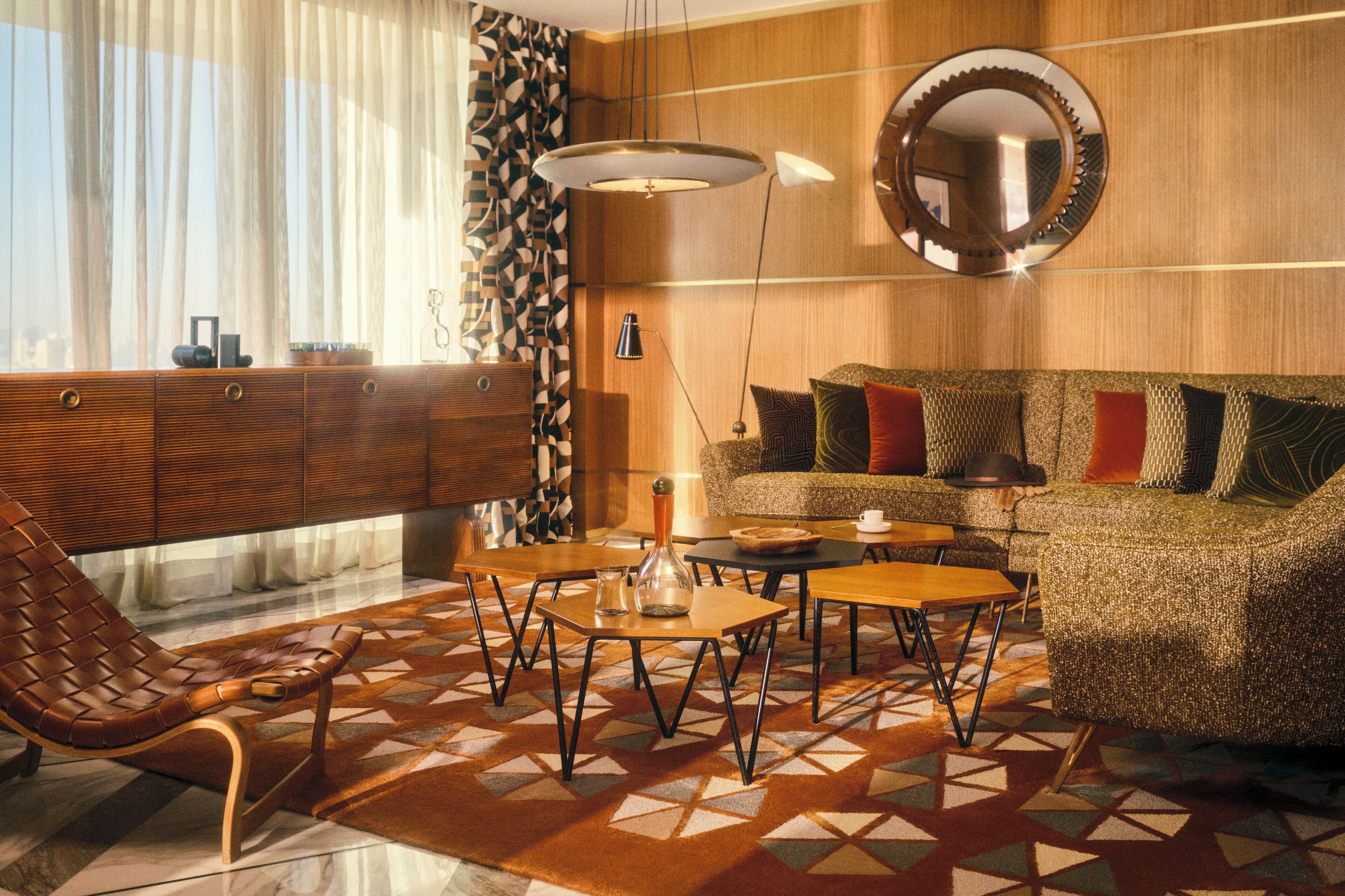 Een moderne woonkamer met een bank, salontafel en een lederen fauteuil. Er hangt een grote spiegel aan de muur en er is een raam met dunne gordijnen.
