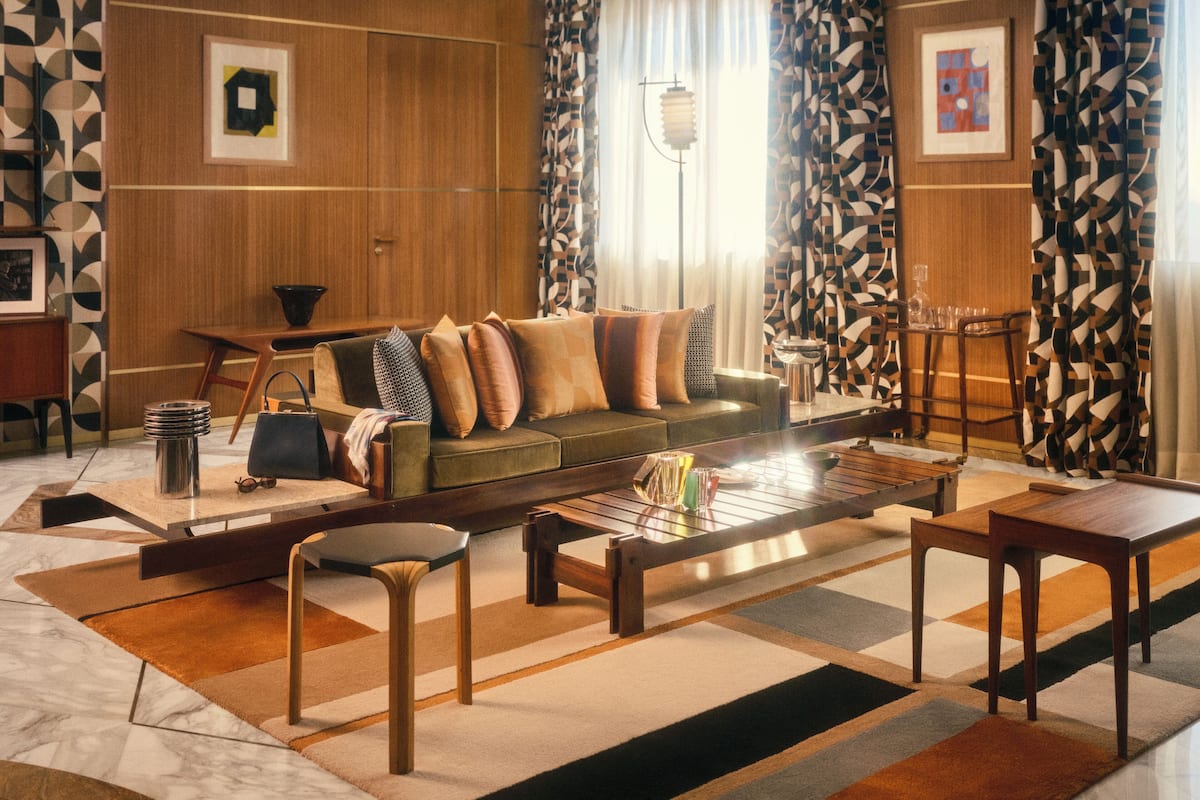 A modern living room with a sofa, coffee table, and chairs. There is a patterned rug on the floor, and a lamp is visible in the background.