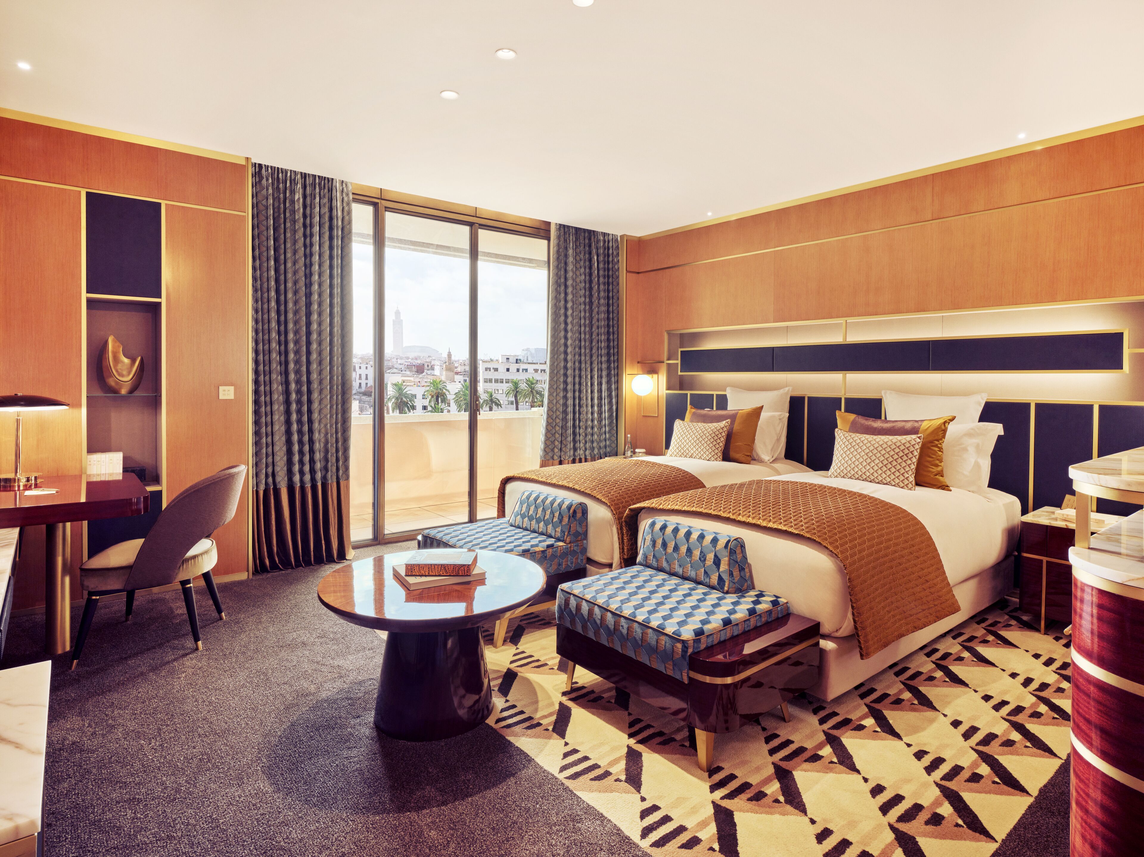 Une chambre d’hôtel avec un grand lit, deux fauteuils, une petite table et une vue sur la ville grâce à de grandes fenêtres.