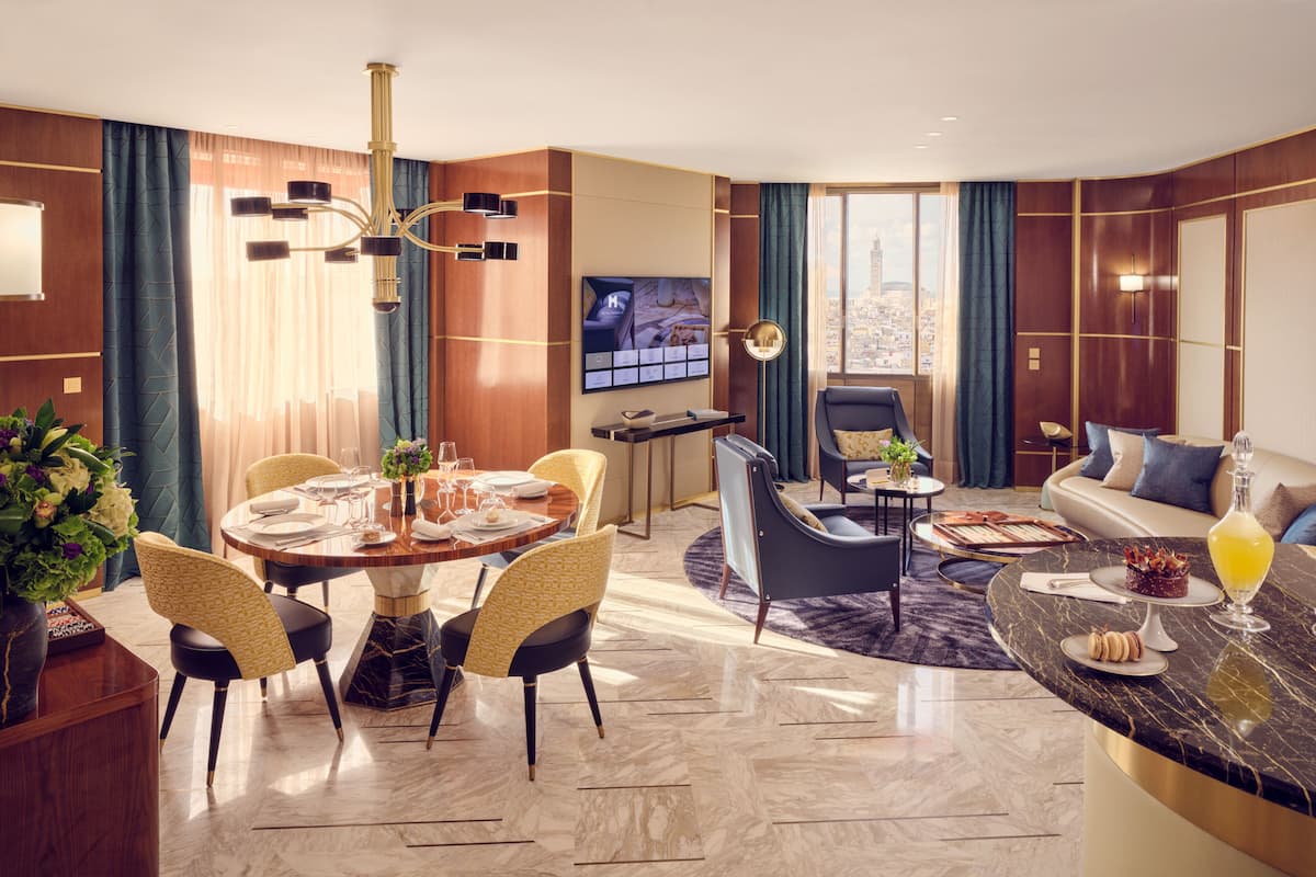 A modern hotel room with a dining area, a living area with a sofa and armchairs, a TV, and a city view.