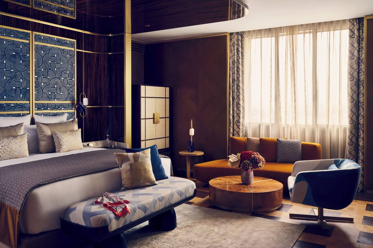 A modern hotel room with a large bed, a sofa, a coffee table, and a chair. There is a window with curtains and a view of the outside.