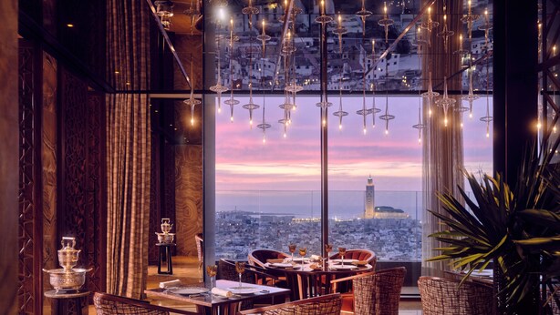Un restaurant situé dans un immeuble de grande hauteur, offrant une vue panoramique sur la ville, doté d’un mobilier élégant et de luminaires suspendus.