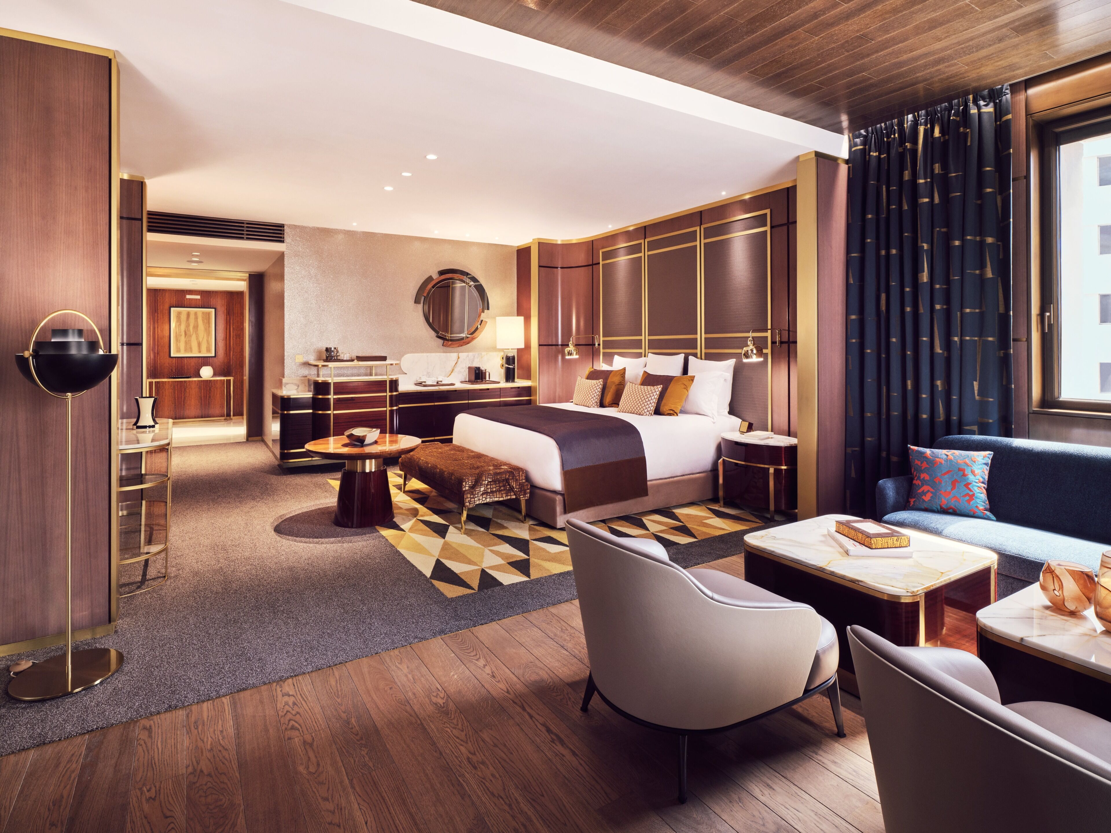 Una habitación de hotel moderna con cama, sofá, sillón, mesa de centro redonda y un escritorio con lámpara.