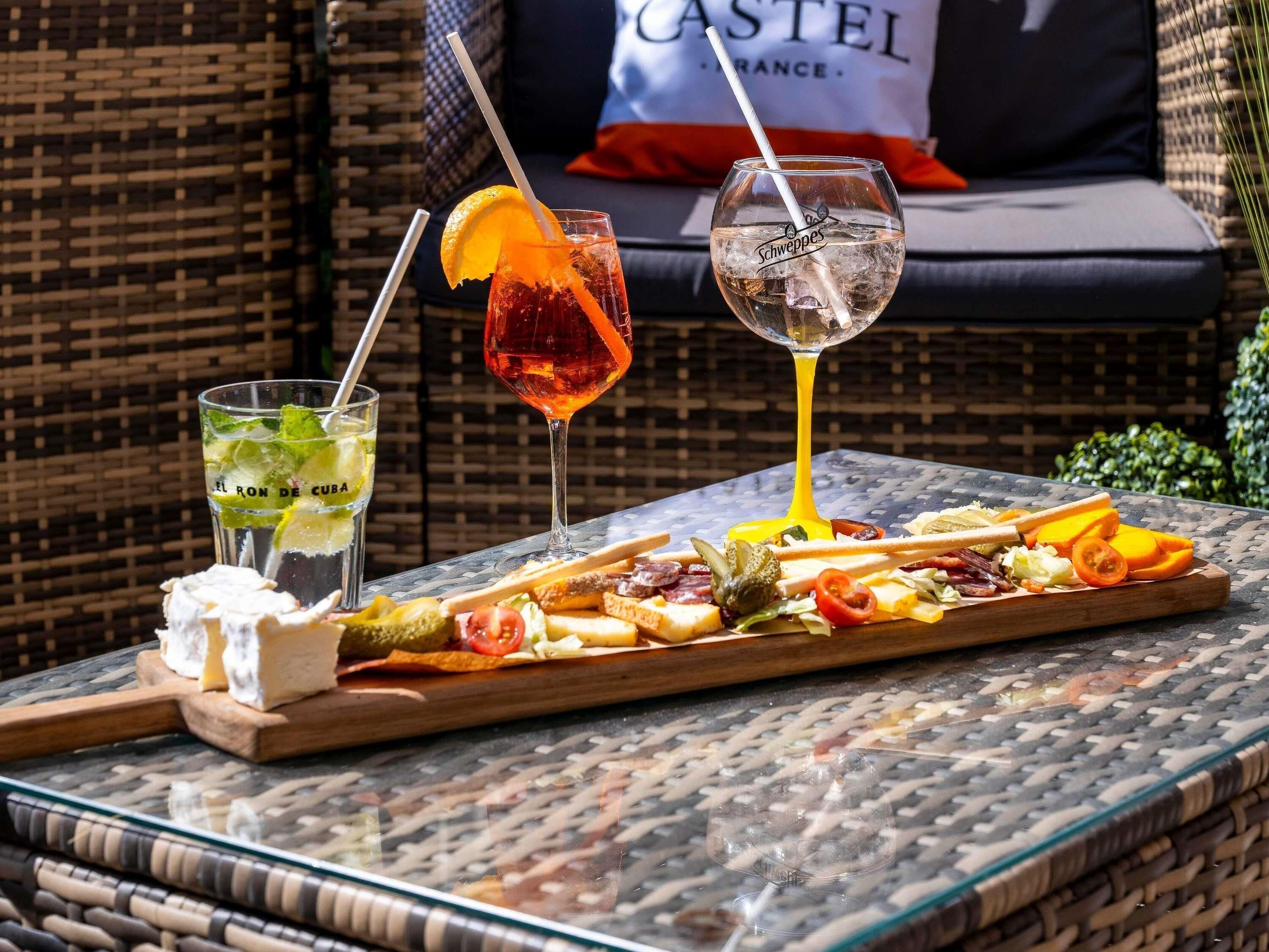 Une table avec une planche de charcuterie, deux boissons et, au fond, une chaise en osier.
