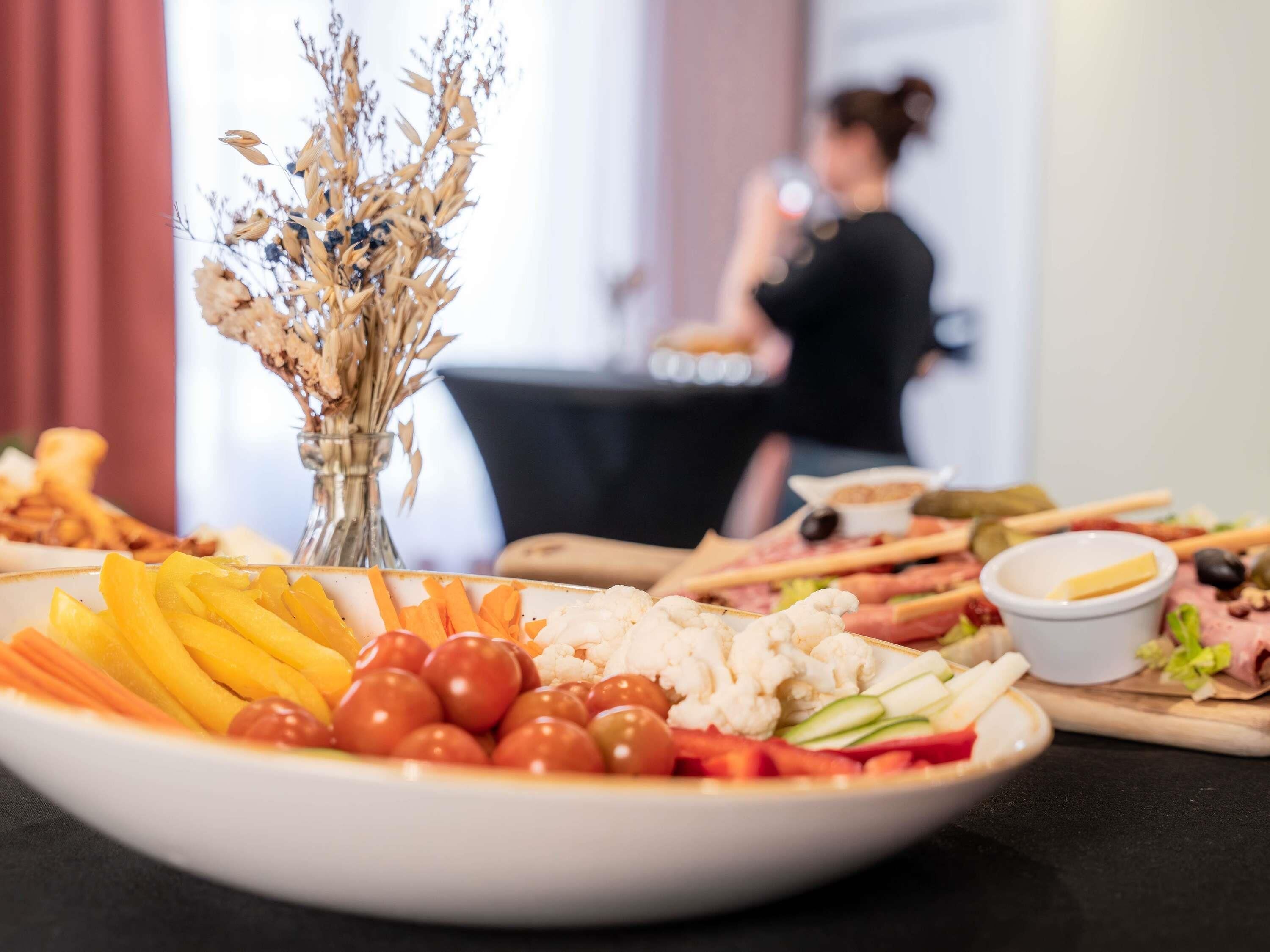 A table with a variety of food items, including fruits, vegetables, and cheese, with a vase of dried flowers in the background.