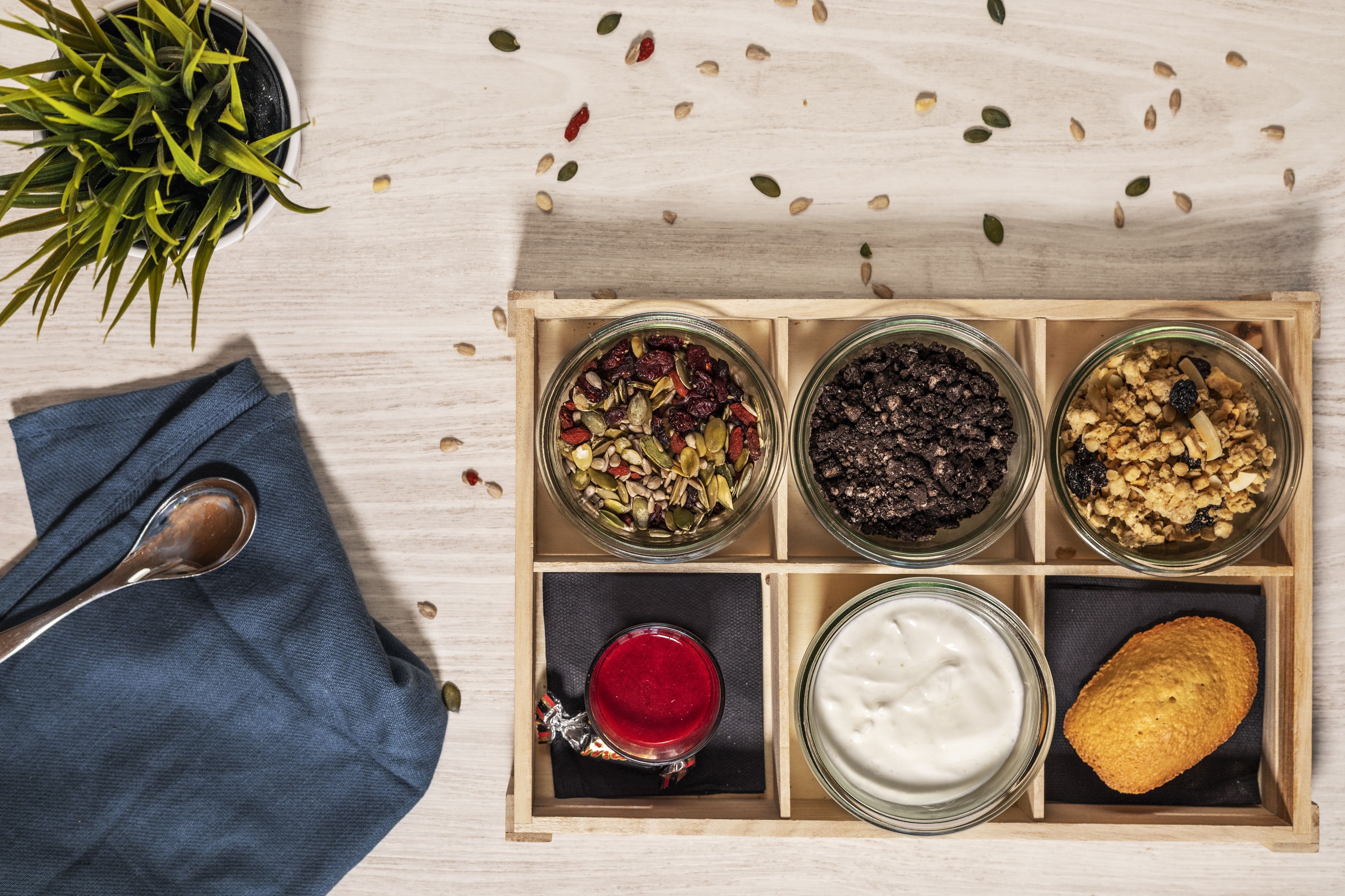 Una bandeja de madera con seis compartimentos que contienen diversos alimentos, una cuchara, un vaso de líquido rojo y una planta.