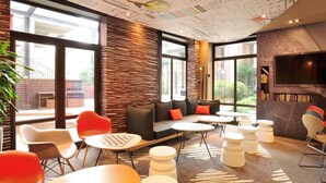 A modern lounge area with a large window, a sofa with orange cushions, and a flat-screen TV mounted on the wall. - Hotel ibis Lille Centre Gares (Lille)