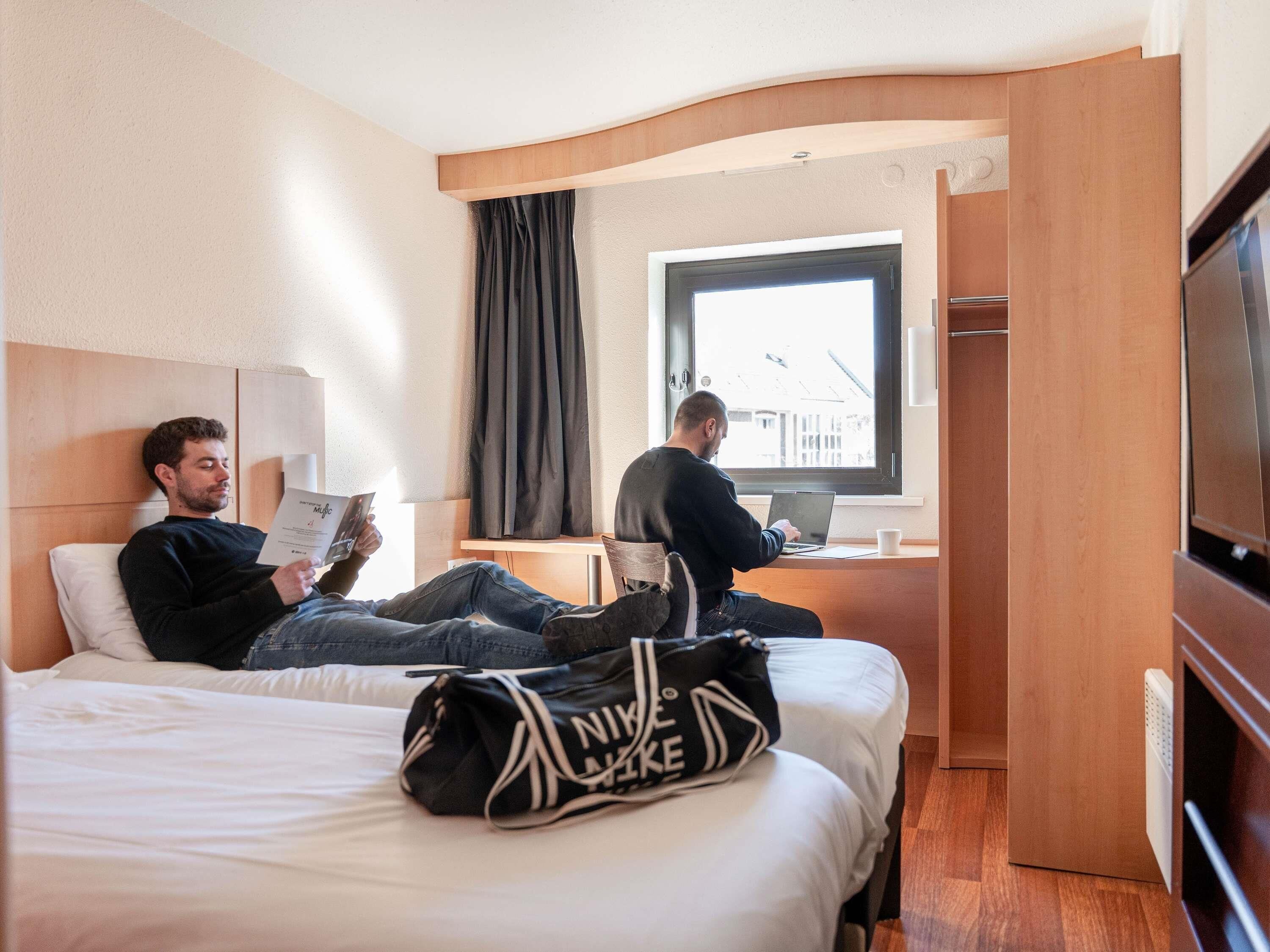 A hotel room with a bed, a desk with a laptop, a window with curtains, and a TV.