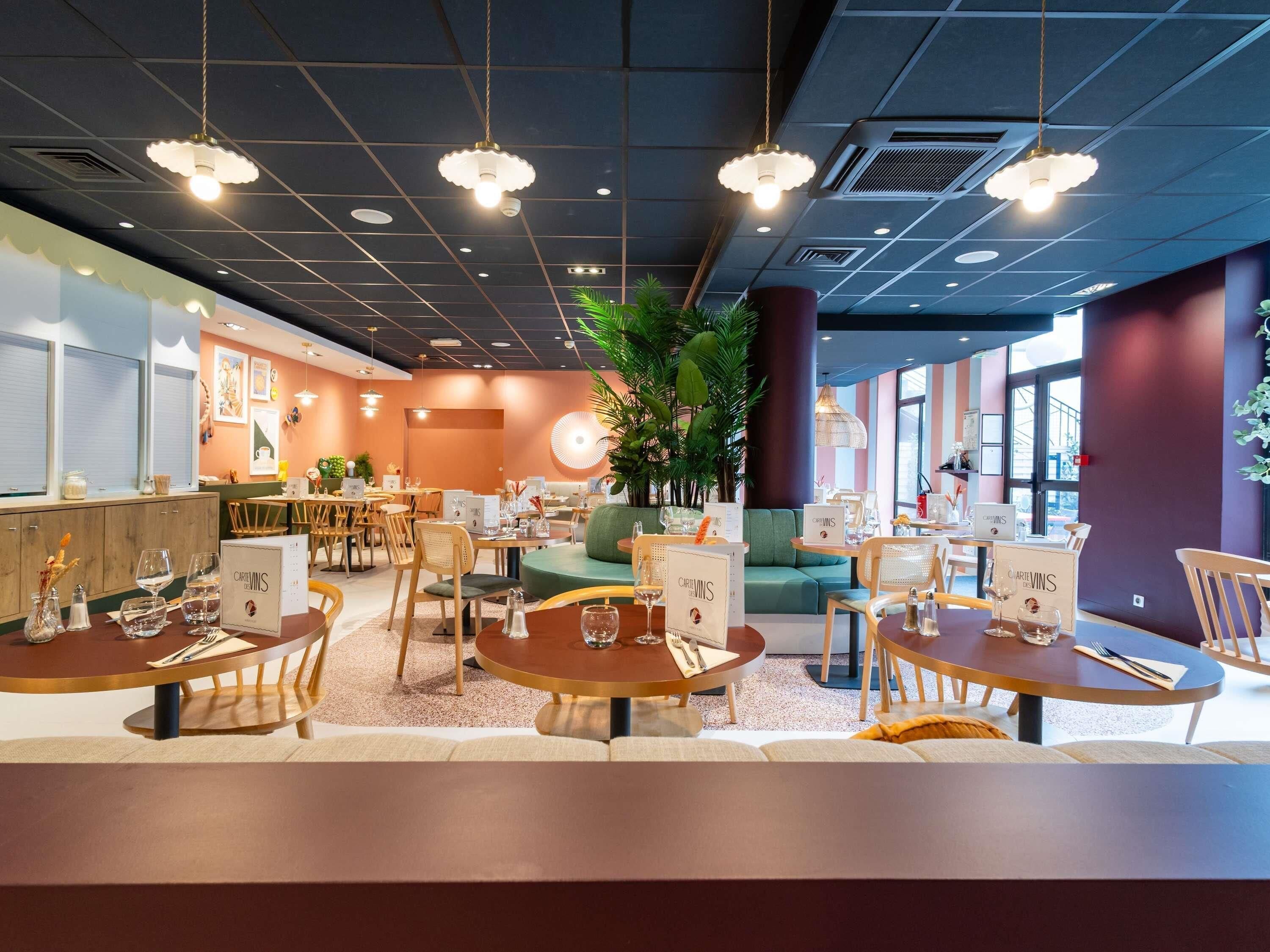 A restaurant interior with round tables, chairs, hanging pendant lights, and a large potted plant.