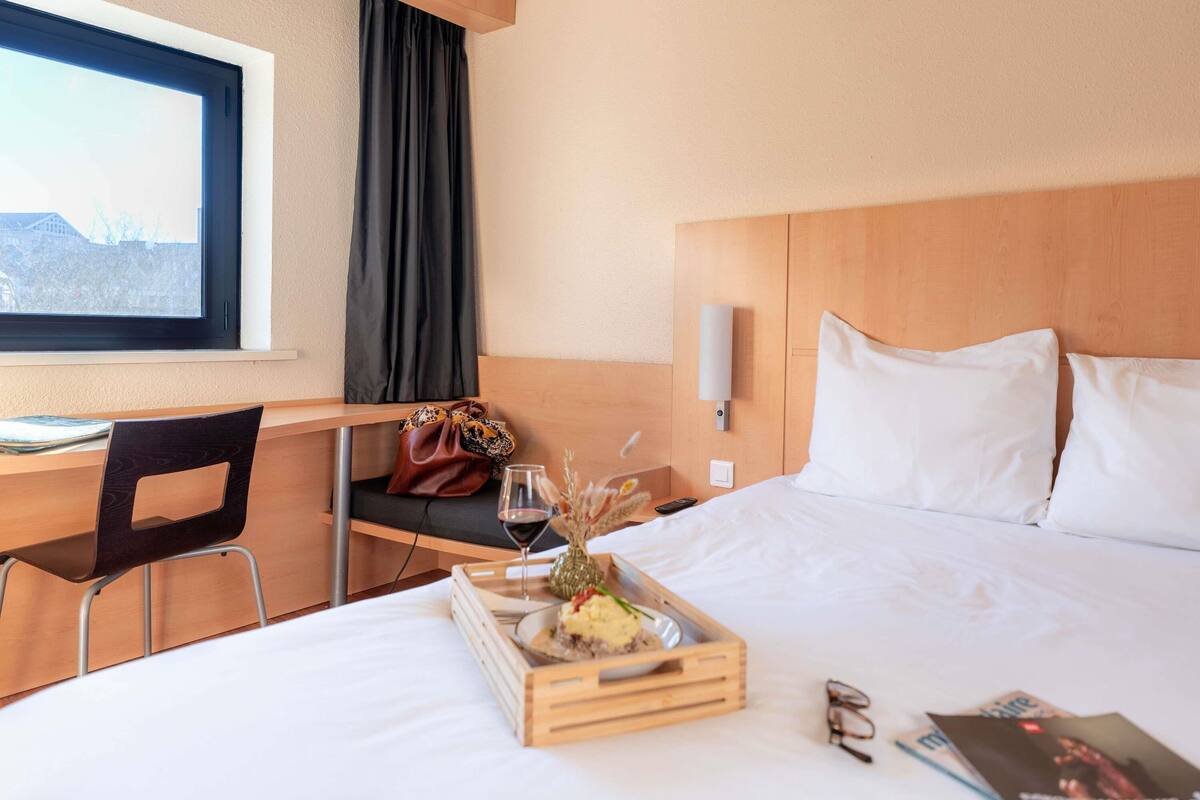 Une chambre d’hôtel comprenant un lit, un bureau, une chaise, une fenêtre avec des rideaux, une table de chevet et un plateau avec de la nourriture et un verre de vin.