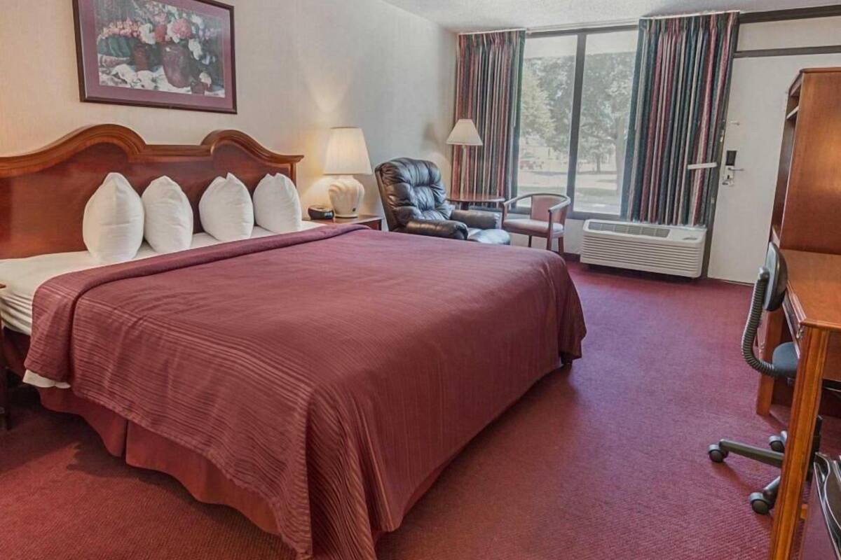 Ein Hotelzimmer mit einem großen Bett, einem Sessel, einem Schreibtisch, einer Lampe, einem Bild und einem Fenster mit Vorhängen.