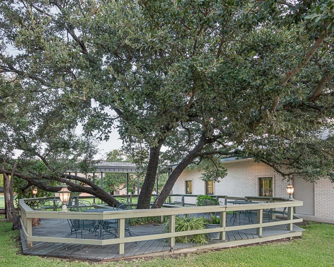 A wooden deck with outdoor seating, surrounded by trees and a building with a brick facade.