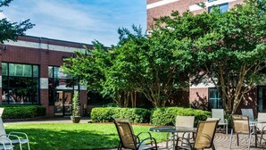 An outdoor seating area with tables and chairs, surrounded by greenery and a building with large windows. - Quality Suites Lake Wright - Norfolk Airport (Norfolk)