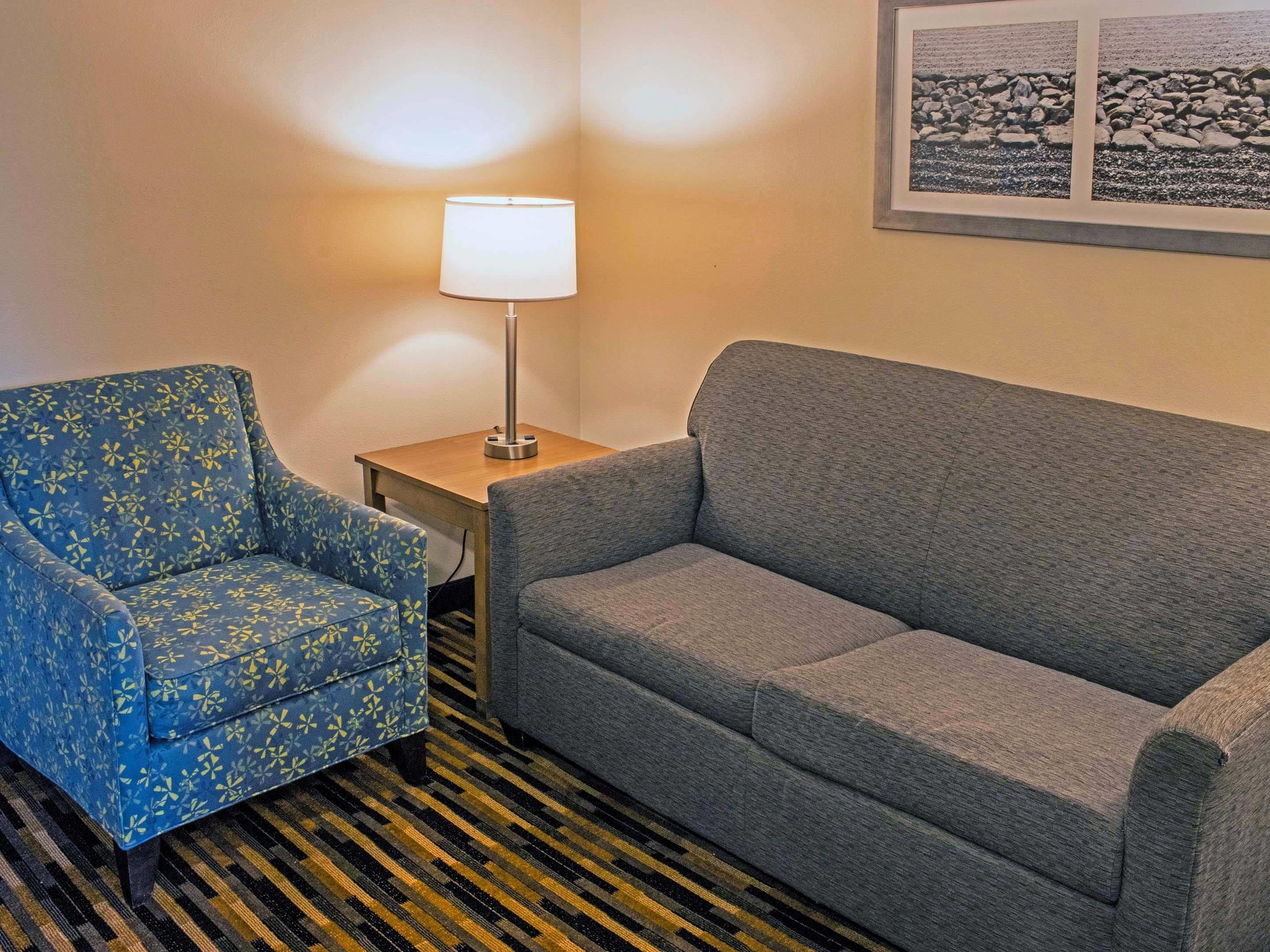 A room with a blue patterned armchair, a grey sofa, a small wooden side table with a lamp, and a framed picture on the wall.