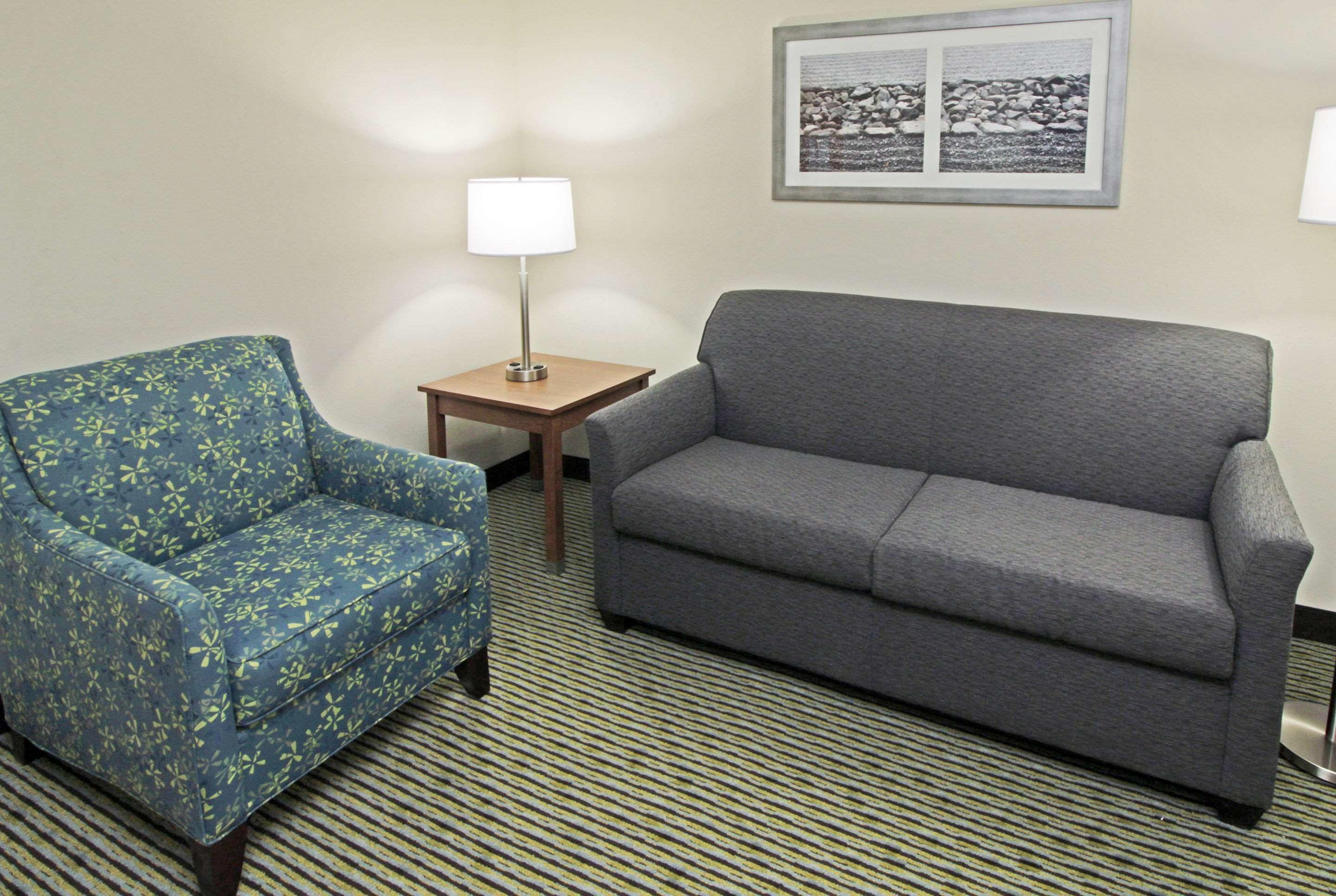 A room with a patterned armchair, a grey sofa, a small table with a lamp, and a framed picture on the wall.