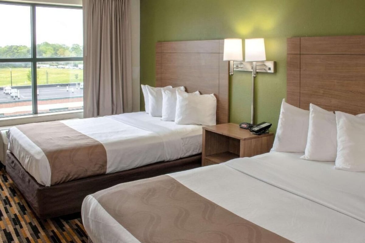 Habitación de hotel con dos camas, una mesita de noche con lámpara y una ventana con vista al aeropuerto.