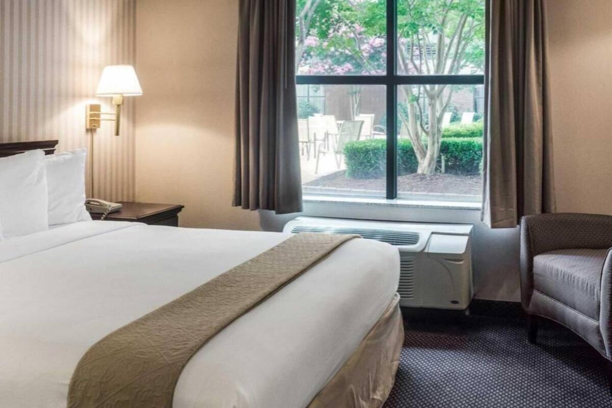 A hotel room with a bed, a desk, a chair, a window with curtains, and a lamp.