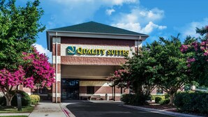 A hotel named Quality Suites with a brick facade, a green roof, and surrounded by lush greenery and pink flowers. - Quality Suites Lake Wright - Norfolk Airport (Norfolk)