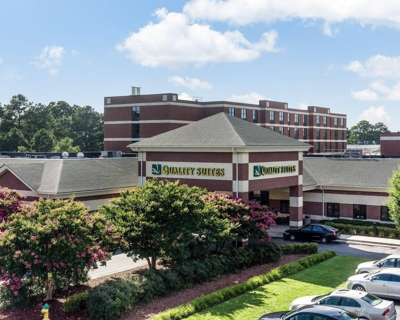 A hotel with a brick facade and multiple windows. There are cars parked in the driveway and a well-maintained lawn area.