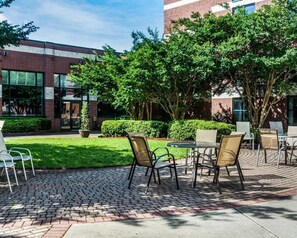 An outdoor patio area with tables and chairs, surrounded by greenery and a building with large windows. - Quality Suites Lake Wright - Norfolk Airport (Norfolk)