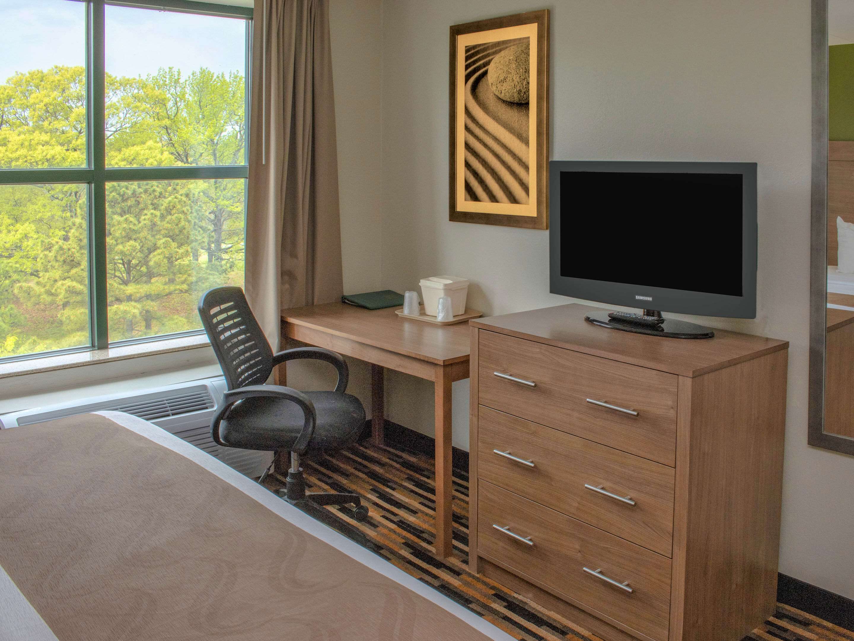 Habitación de hotel con cama, escritorio con computadora, silla, cómoda y ventana con vista a árboles.