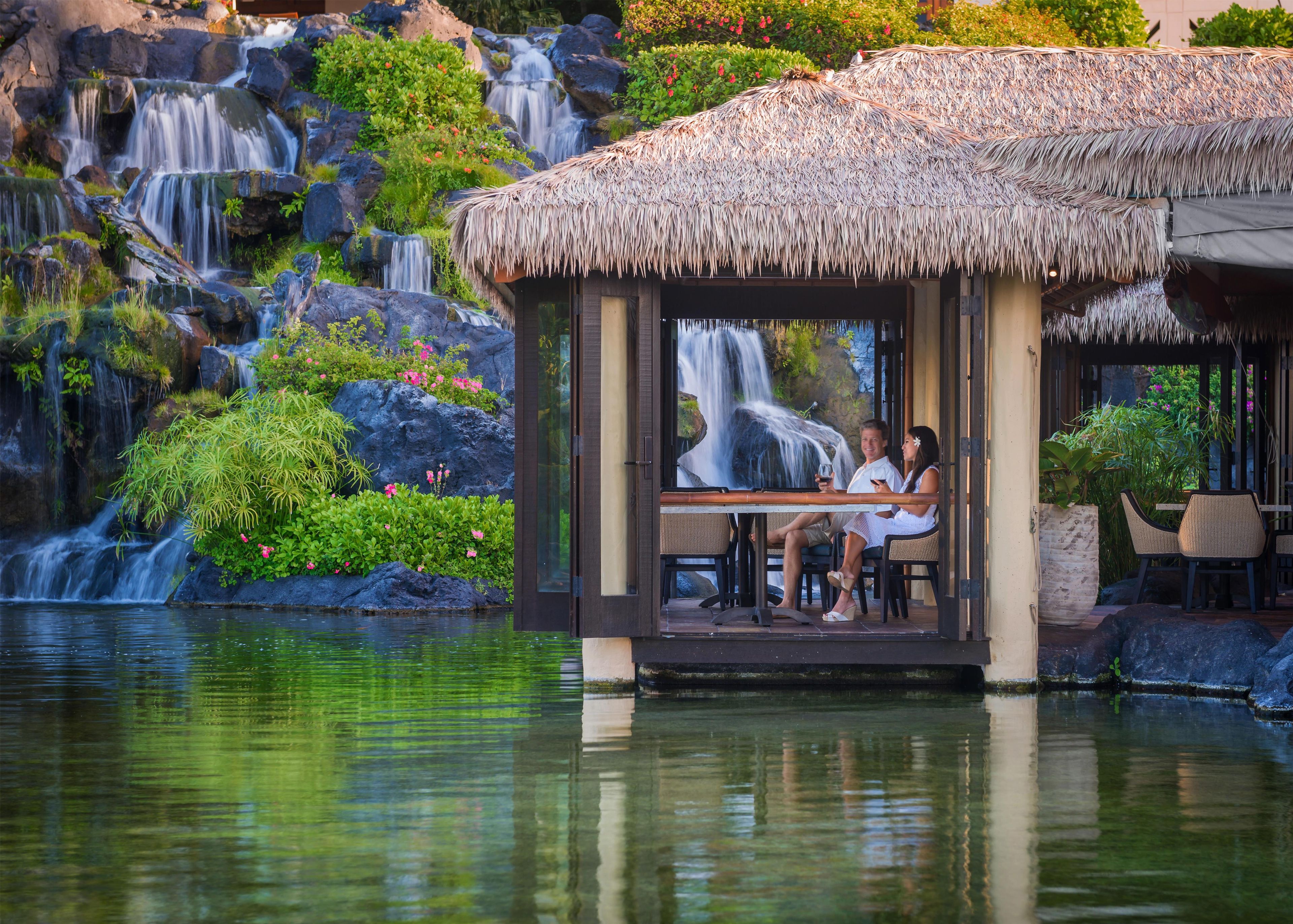 Estructura de techo de paja con vistas a una cascada, un área de comedor con sillas y una superficie acuática reflectante.