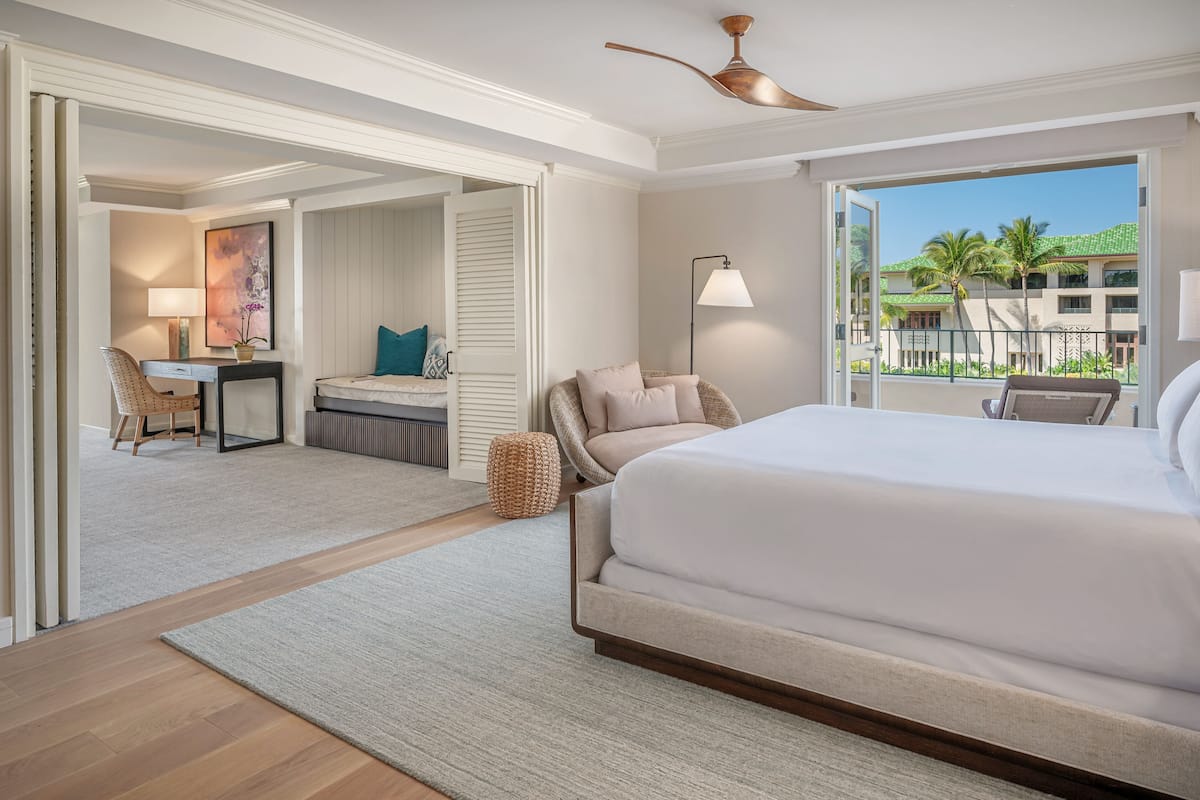 A spacious bedroom with a large bed, a desk, and a chair, a balcony with a view of palm trees, and a ceiling fan.