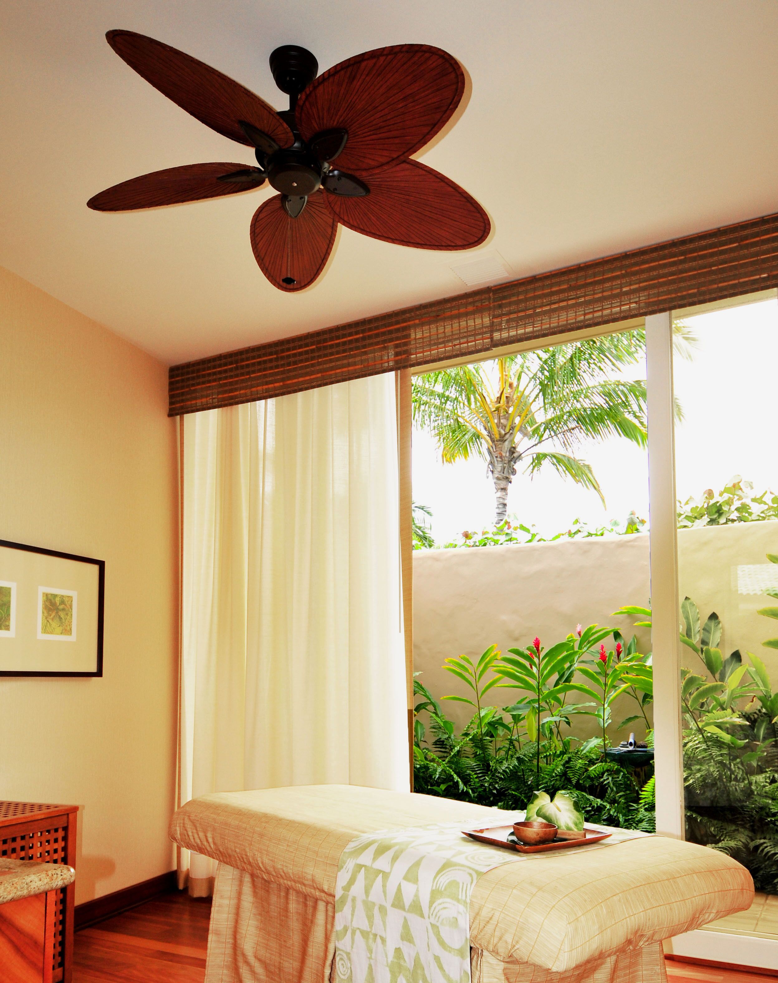 A room with a bed, a ceiling fan, a window with curtains, and a view of greenery outside.
