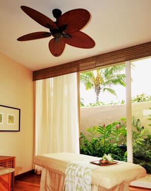 A room with a bed, a ceiling fan, a window with curtains, and a view of greenery outside.