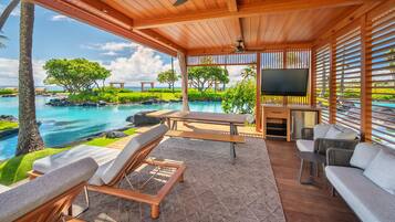 A covered outdoor seating area with a pool view, wooden furniture, and a flat-screen TV.