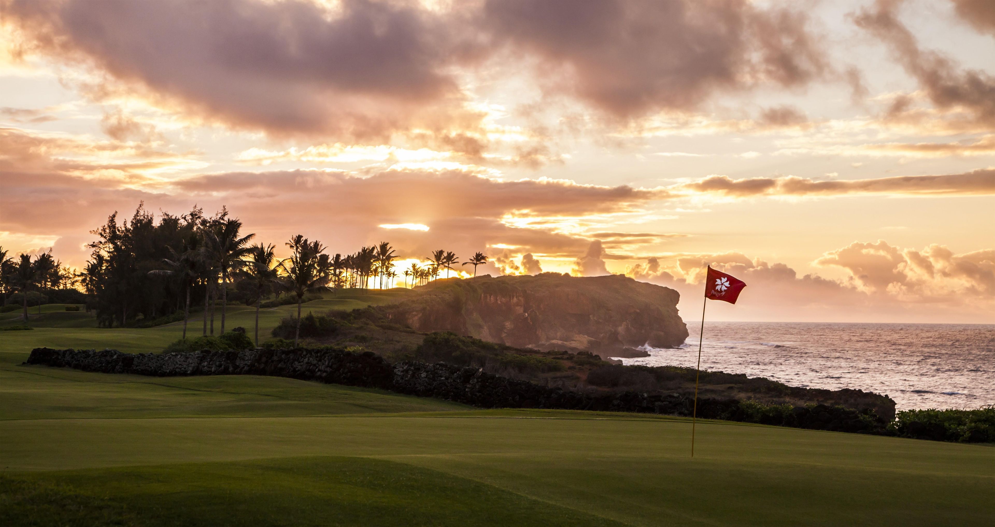 Campo de golf con una bandera, palmeras y un acantilado con vista al océano al atardecer.