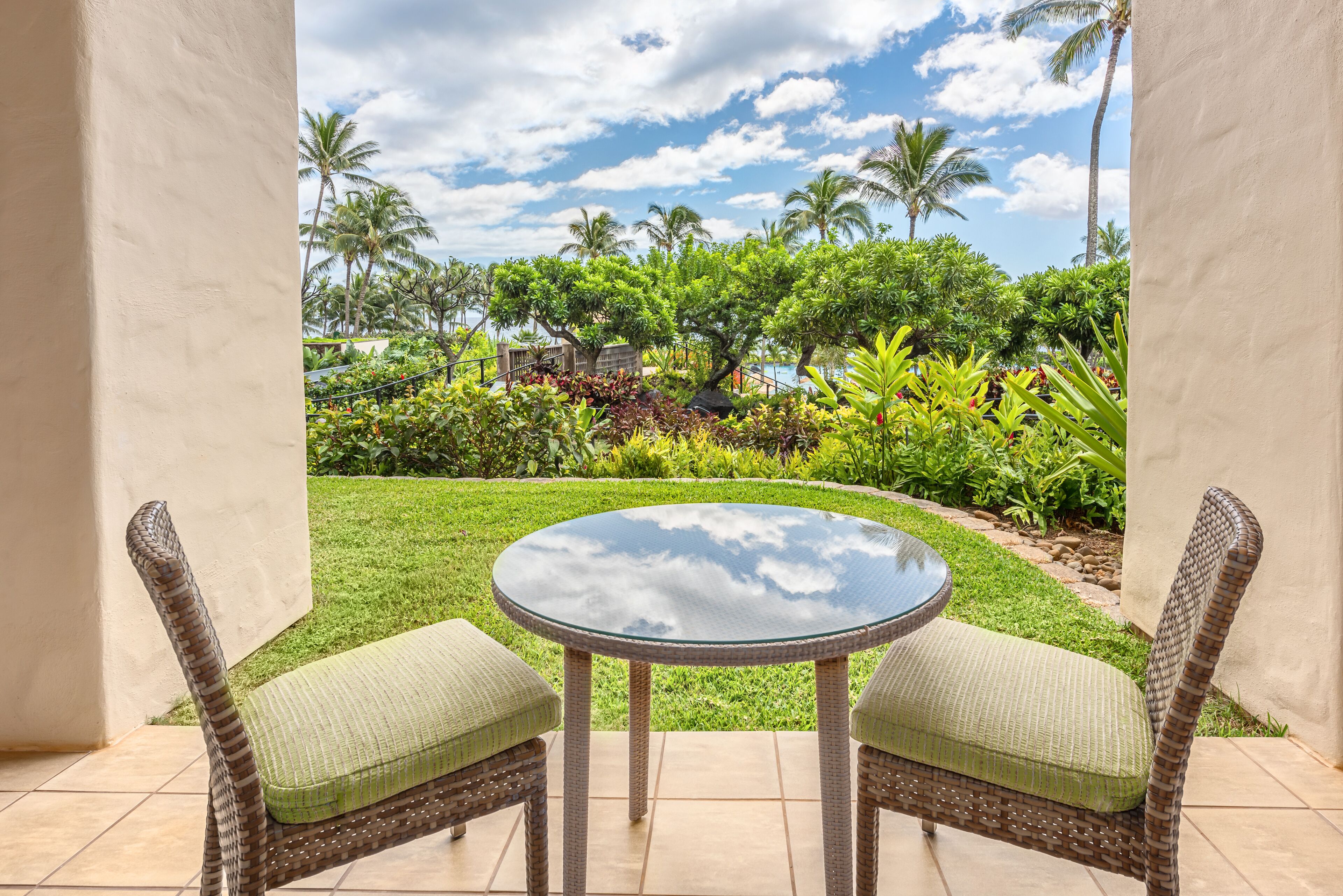 Une terrasse dotée de chaises en osier et d’une table en verre, offrant une vue sur un jardin luxuriant, avec des palmiers et un ciel dégagé.