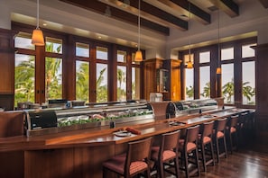 A bar area with a long counter, multiple chairs, and a view of palm trees through large windows.