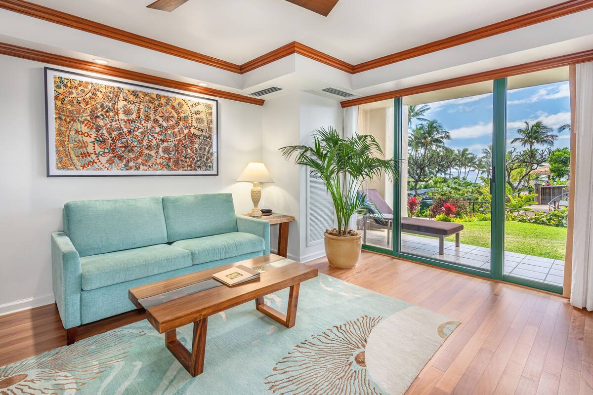 Un salon avec un canapé bleu clair, une table basse en bois, un grand tableau abstrait, une plante en pot et une vue sur un jardin tropical, visible à travers des portes vitrées.