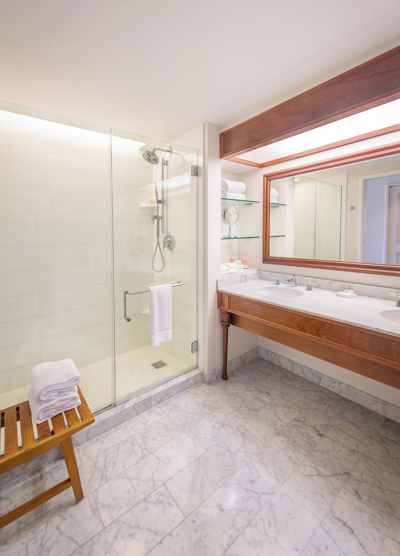 A bathroom with a glass-enclosed shower, a wooden bench with towels, and a large mirror above a marble countertop.